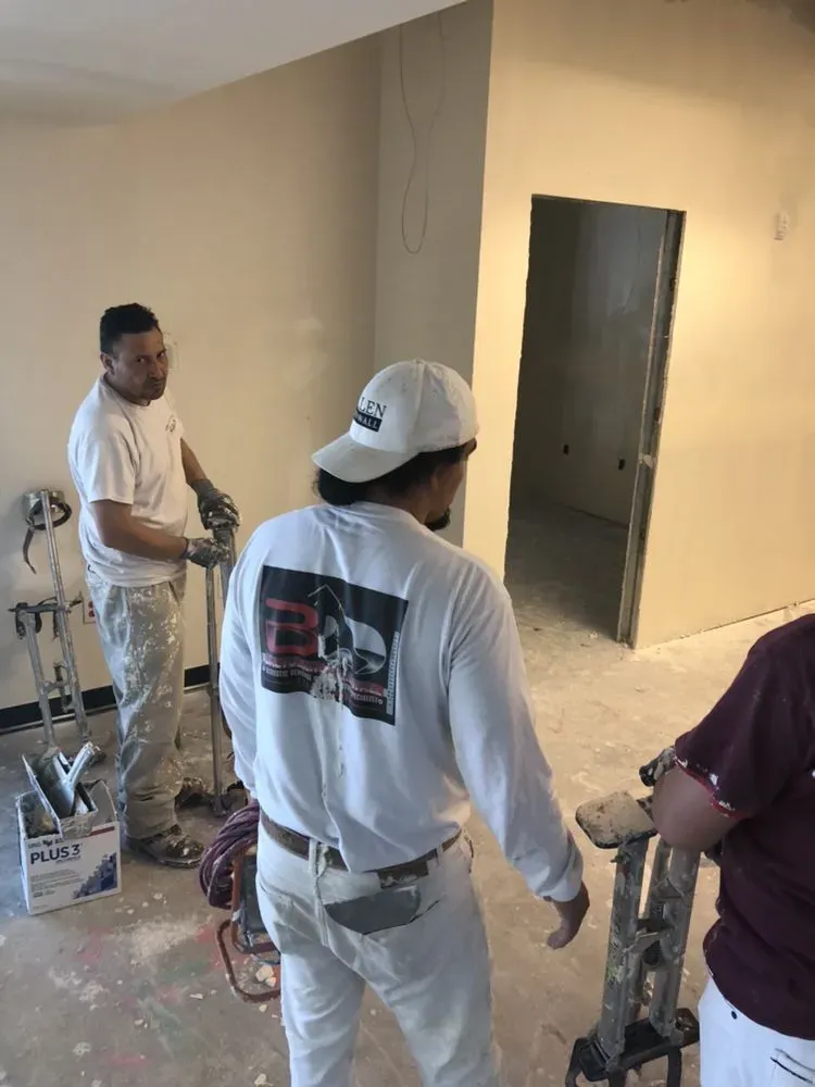 "Three workers from Baltodano's Drywall and Painting stand in a room under renovation, wearing work attire and surrounded by drywall tools and materials."